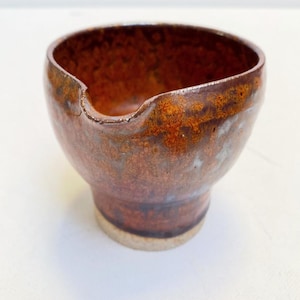 Op de afbeelding: Een handgemaakte keramische kom met een tuit, met een mix van bruine en oranje tinten. De kom heeft een afgeronde vorm met een licht uitlopende basis en een getextureerd oppervlak. De binnenkant is donkerder bruin en de buitenkant heeft een glanzende afwerking.