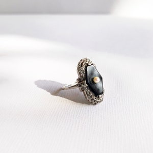 May include: A silver ring with a black onyx stone and a small pearl accent. The ring has a detailed filigree design.