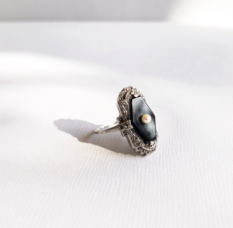 May include: A silver ring with a black onyx stone and a small pearl accent. The ring has a detailed filigree design.