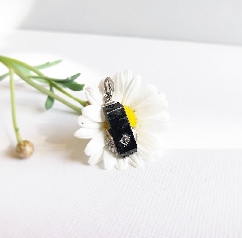 May include: A black gemstone pendant with a silver setting and a small diamond accent. The pendant is resting on a white daisy.