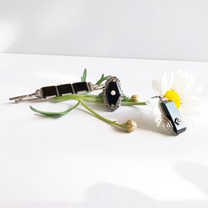 May include: Three black onyx jewelry pieces on a white background with a white daisy. The pieces are a brooch, a ring, and a pendant.