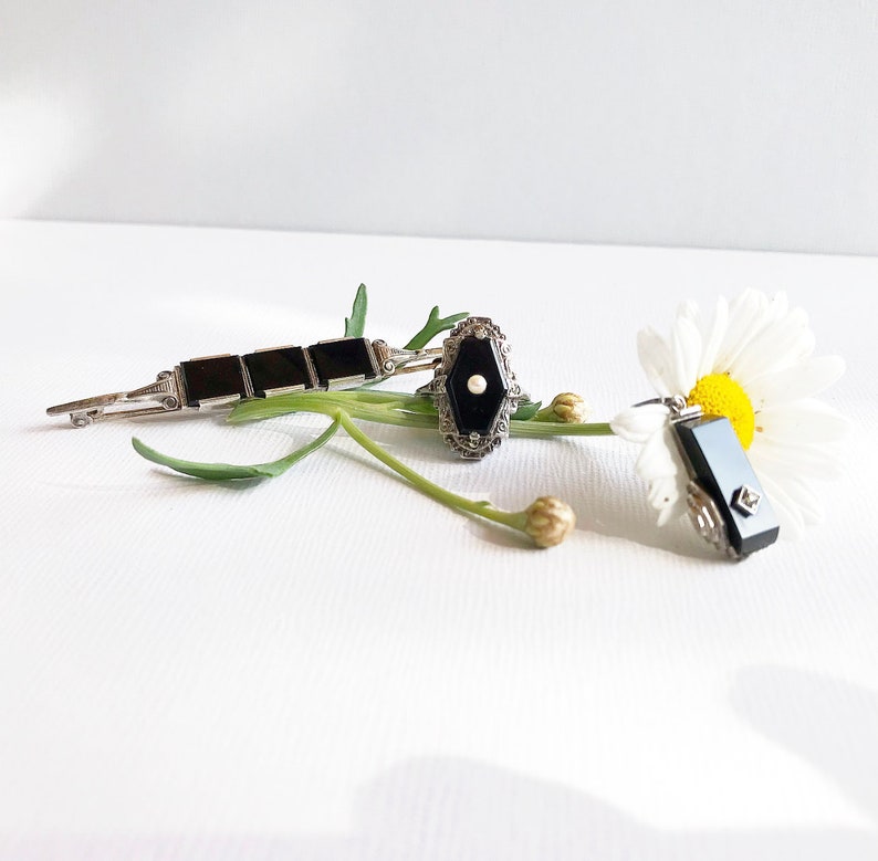May include: Three black onyx jewelry pieces on a white background with a white daisy. The pieces are a brooch, a ring, and a pendant.