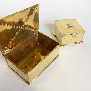 Pode incluir: Duas caixas de lat&atilde;o, uma com tampa articulada e a outra com tampa quadrada. A caixa com a tampa articulada est&aacute; aberta, revelando o interior vazio. Ambas as caixas t&ecirc;m detalhes decorativos e s&atilde;o feitas de um metal brilhante de cor dourada.