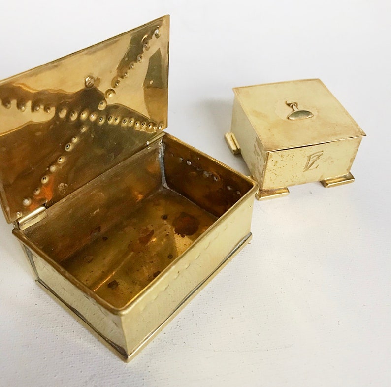 Pode incluir: Duas caixas de lat&atilde;o, uma com tampa articulada e a outra com tampa quadrada. A caixa com a tampa articulada est&aacute; aberta, revelando o interior vazio. Ambas as caixas t&ecirc;m detalhes decorativos e s&atilde;o feitas de um metal brilhante de cor dourada.
