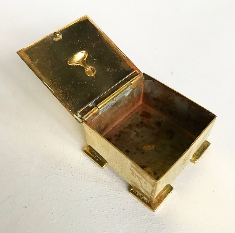 Pode incluir: Uma pequena caixa quadrada de lat&atilde;o com uma tampa articulada. A caixa tem uma pequena pega redonda de lat&atilde;o na tampa. O interior da caixa est&aacute; vazio.