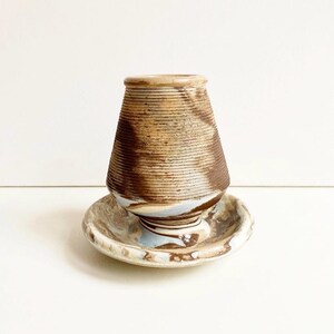 Peut inclure: Un vase en céramique fait à la main avec un design texturé et nervuré dans des tons bruns et beiges. Le vase repose sur une soucoupe assortie avec un effet marbré, avec des touches de bleu et de brun. Une pièce décorative unique.