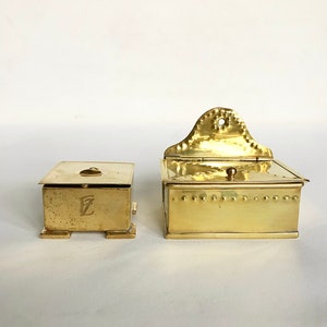 Pode incluir: Duas caixas antigas de lat&atilde;o com tampas articuladas. A caixa menor tem uma letra "E" em relevo na frente. A caixa maior tem um design decorativo na tampa.
