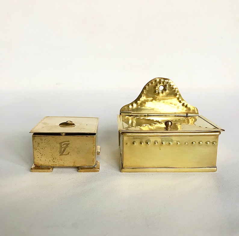Pode incluir: Duas caixas antigas de lat&atilde;o com tampas articuladas. A caixa menor tem uma letra "E" em relevo na frente. A caixa maior tem um design decorativo na tampa.
