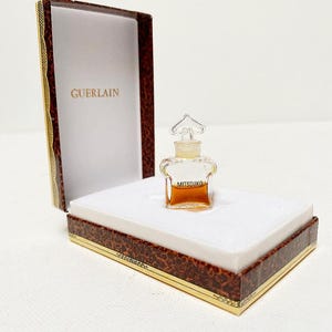 Könnte beinhalten: Eine kleine, durchsichtige Glasflasche Mitsouko-Parfüm von Guerlain in der Originalverpackung. Die Flasche hat einen goldfarbenen Verschluss und die Schachtel ist braun mit goldenen Verzierungen.