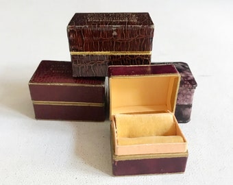 Pode incluir: Quatro pequenas caixas de joias vintage. As caixas são bordô com detalhes dourados e uma aparência de couro sintético texturizado. Uma caixa está aberta, revelando um interior amarelo com uma almofada de veludo. As caixas estão dispostas sobre uma superfície branca.