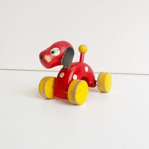 Può includere: Un giocattolo vintage in legno a forma di cane da trascinare. Il cane è dipinto di rosso con pois bianchi, ruote gialle e una palla gialla sulla schiena. Il cane ha orecchie nere e un occhio verde.