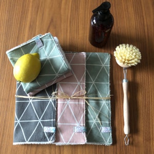 Könnte beinhalten: Drei wiederverwendbare Reinigungstücher mit geometrischem Muster in Grün, Rosa und Grau. Die Tücher sind mit einem natürlichen Bindfadenbogen zusammengebunden. Eine Zitrone, eine Flasche Reinigungslösung und eine Holzschrubbbürste sind ebenfalls im Bild.