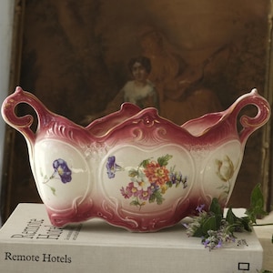 Könnte beinhalten: Eine längliche Keramikschale als Tafelaufsatz mit einem Farbverlauf in Dunkelrosa und Weiß sowie verzierten, geschwungenen Griffen. Die Außenseite ist mit handgemalten Blumenmotiven verziert, darunter blaue Prunkwinden und ein zentraler Blumenstrauß.