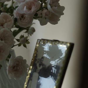 Könnte beinhalten: Ein rechteckiger Bilderrahmen mit einem metallisch goldenen Rand, der ein schwarz-weißes Foto einer Person enthält. Der Rahmen steht neben einem Strauß blassrosa Rosen mit grünen Stielen und Blättern, die sich im Glas des Rahmens spiegeln.