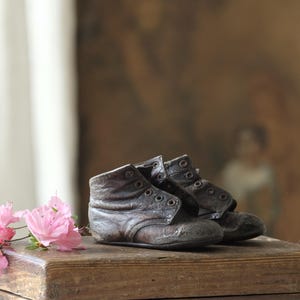 Könnte beinhalten: Ein Paar antike, abgenutzte Baby-Lederstiefel mit Ösen, die auf einer verwitterten Holzkiste liegen. Rosa Blüten befinden sich im Vordergrund, mit einem verschwommenen Hintergrund im Vintage-Stil.