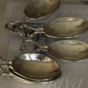 Könnte beinhalten: Ein Set aus vier silberfarbenen Metalllöffeln mit verzierten Griffen, die komplizierte Schnörkel und detaillierte Muster aufweisen. Die Löffelblätter haben eine polierte, reflektierende Oberfläche mit sichtbaren Anzeichen von natürlicher Patina.