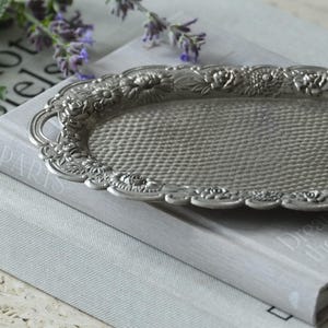 Könnte beinhalten: Ein ovales Tablett aus silberfarbenem Metall mit einer texturierten, genoppten Mitteloberfläche und einem erhöhten Rand, der mit einem detaillierten Reliefmuster aus verschiedenen Blumen und Blättern, wie Rosen und Gänseblümchen, verziert ist.