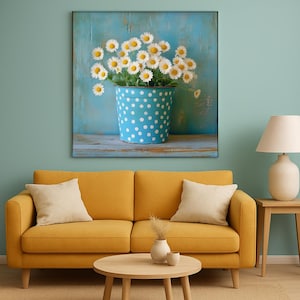 Op de afbeelding: Een canvas print met een blauwe emmer met witte stippen, gevuld met witte madeliefjes. De emmer staat op een houten oppervlak tegen een blauwe achtergrond. De kamer heeft een gele bank en een houten tafel.