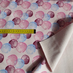 Könnte beinhalten: Stoff mit einem sich wiederholenden Muster aus rosa, blauen und burgunderfarbenen Kreisen auf weißem Hintergrund. Ein gelbes Maßband liegt auf dem Stoff. Der Stoff ist gefaltet und zeigt eine schlichte, hellrosa, strukturierte Unterseite.