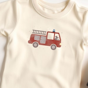 Könnte beinhalten: Ein cremefarbenes T-Shirt mit einer roten Feuerwehrwagen-Illustration. Der Feuerwehrwagen hat eine Leiter, ein blaues Licht und eine kleine Maus, die ihn fährt.