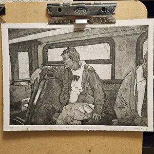 Peut inclure: Une gravure en noir et blanc de deux personnes assises dans un bus. La personne au premier plan porte une veste et une chemise blanche avec une lettre "W" dessus. La personne à l'arrière-plan porte un sweat à capuche.