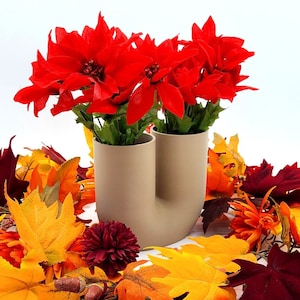 May include: A beige ceramic vase with two openings holds a bouquet of red poinsettia flowers. The vase is surrounded by a wreath of fall leaves in shades of orange, yellow, and red.
