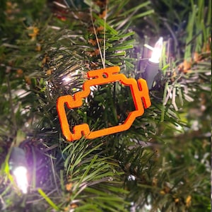 May include: Orange Christmas tree decoration in the shape of a car engine warning light. The ornament hangs on a green Christmas tree with white lights. The decoration is made of a textured material.
