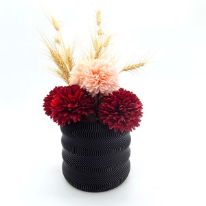 May include: A black wavy vase with a bouquet of pink and red carnations and dried wheat.