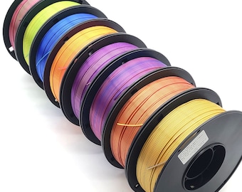 May include: A set of seven spools of filament for 3D printing. The spools are arranged in a stack, with the colors of the filament progressing from red to gold. The spools are all black and have a label on the side.