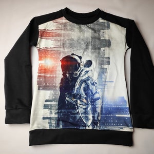 Könnte beinhalten: Schwarzes Langarmshirt mit einem weißen Vorderteil, das eine digitale Stadtlandschaft und einen Astronauten in einem Raumanzug zeigt. Der Astronaut steht vor einem leuchtend orangenen Licht.