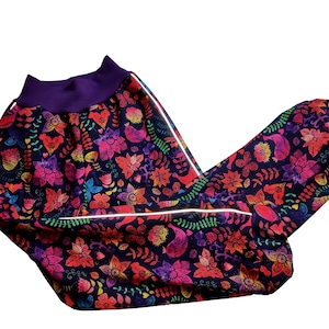 Könnte beinhalten: Eine farbenfrohe Hose mit Blumen- und Vogelmuster. Die Hose hat einen lila Bund und reflektierende Paspeln entlang der Nähte.