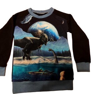 Può includere: Una felpa nera a maniche lunghe con una grafica a tema dinosauri e spazio. Il design presenta un T-Rex, un dinosauro dal collo lungo, pianeti e un sottomarino in un paesaggio blu e verde. I polsini e lo scollo sono azzurri.