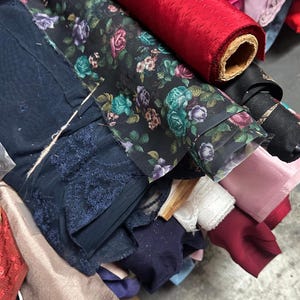 Puede incluir: Una variedad de rollos de tela en diferentes colores y patrones, incluyendo florales, de encaje y colores s&oacute;lidos. Los rollos est&aacute;n apilados uno encima del otro, creando una exhibici&oacute;n colorida y texturizada.