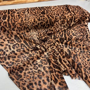 K&ouml;nnte beinhalten: Ein gefaltetes St&uuml;ck transparenten Stoffes mit Leopardenmuster. Der Stoff hat einen beigefarbenen Hintergrund mit dunkelbraunen Flecken, wodurch ein klassisches Tiermuster entsteht. Der Stoff wird wahrscheinlich f&uuml;r Kleidung oder Dekorationszwecke verwendet.