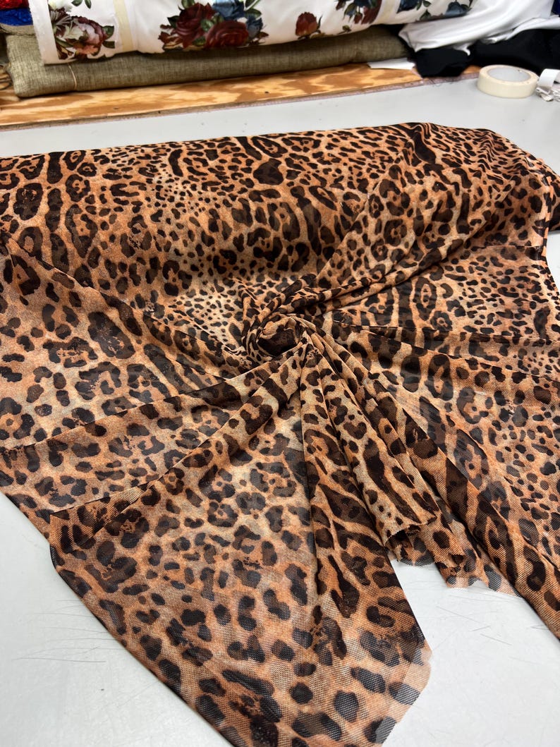 K&ouml;nnte beinhalten: Ein gefaltetes St&uuml;ck transparenten Stoffes mit Leopardenmuster. Der Stoff hat einen beigefarbenen Hintergrund mit dunkelbraunen Flecken, wodurch ein klassisches Tiermuster entsteht. Der Stoff wird wahrscheinlich f&uuml;r Kleidung oder Dekorationszwecke verwendet.