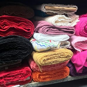 Puede incluir: Un estante lleno de rollos de tela de varios colores, incluyendo azul, verde, rojo, negro, rosa y blanco. La tela est&aacute; cuidadosamente apilada y organizada.