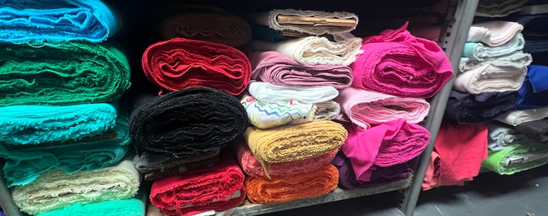 Puede incluir: Un estante lleno de rollos de tela de varios colores, incluyendo azul, verde, rojo, negro, rosa y blanco. La tela est&aacute; cuidadosamente apilada y organizada.