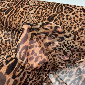 K&ouml;nnte beinhalten: Eine Rolle transparenten Stoffes mit Leopardenmuster. Der Stoff hat einen beigefarbenen Hintergrund mit braunen und schwarzen Flecken. Das Material ist leicht und scheint f&uuml;r Kleidung oder Dekorationszwecke geeignet zu sein.