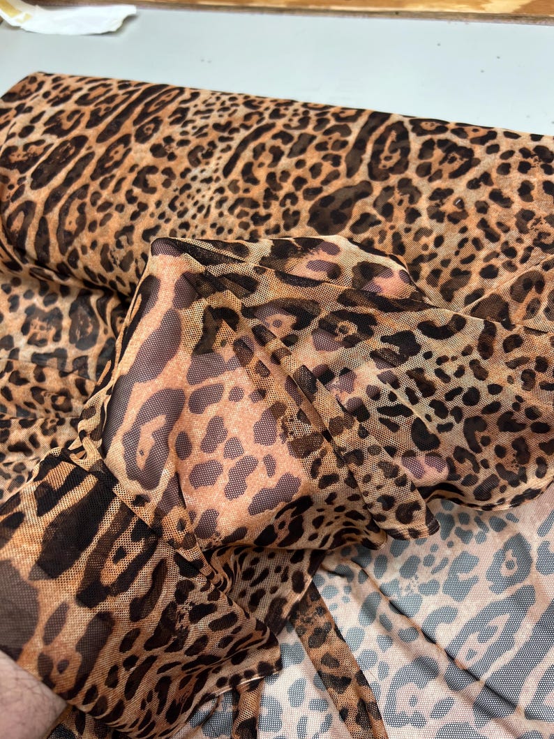 K&ouml;nnte beinhalten: Eine Rolle transparenten Stoffes mit Leopardenmuster. Der Stoff hat einen beigefarbenen Hintergrund mit braunen und schwarzen Flecken. Das Material ist leicht und scheint f&uuml;r Kleidung oder Dekorationszwecke geeignet zu sein.