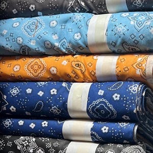 May include: A stack of colorful fabric with bandana patterns. The fabric is rolled up and has a white label on each roll. The colors include blue, yellow, orange, black, and red.