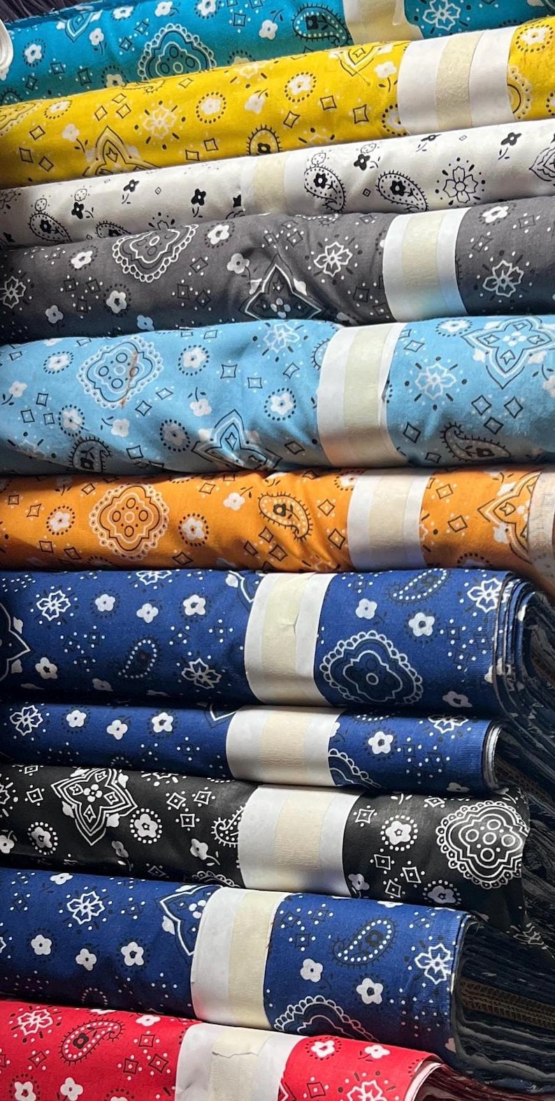 May include: A stack of colorful fabric with bandana patterns. The fabric is rolled up and has a white label on each roll. The colors include blue, yellow, orange, black, and red.