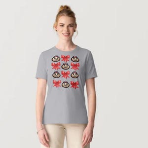 May include: A light gray short-sleeved t-shirt featuring a repeating pattern of small cartoon faces wearing dark caps and bright red decorative bows arranged in a grid across the center of the chest area of the garment.