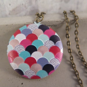 Könnte beinhalten: Eine runde Anhänger-Kette mit einem bunten Muster aus überlappenden Schuppen in den Farben Rosa, Rot, Blau, Weiß und Schwarz. Die Kette ist bronzefarben.