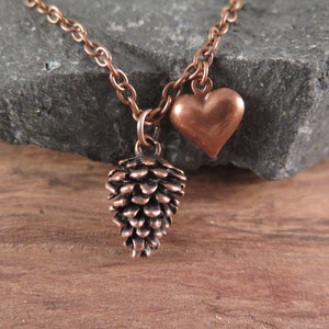Könnte beinhalten: Eine kupferne Halskette mit einem Herz- und einem Tannenzapfen-Anhänger. Die Kette liegt auf einem dunkelgrauen Stein und einer Holzoberfläche.
