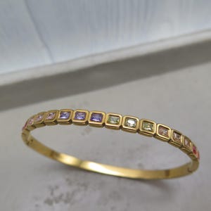 Peut inclure: Un bracelet jonc doré orné de pierres carrées multicolores, notamment roses, violettes et vertes. Le bracelet incurvé est mis en valeur sur un fond neutre, soulignant les pierres précieuses colorées.