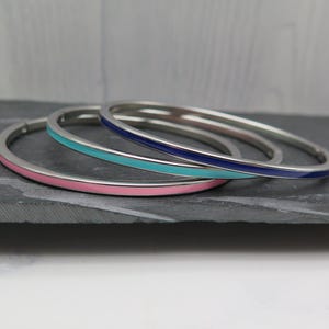 Könnte beinhalten: Drei silberne Armreifen mit Emaillebändern in Rosa, Türkis und Marineblau. Die Armbänder werden auf einer dunkelgrauen Oberfläche mit einem hellgrauen Hintergrund präsentiert.