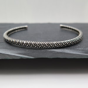 Könnte beinhalten: Ein silbernes geflochtenes Armreif-Armband mit einem schlichten, eleganten Design.