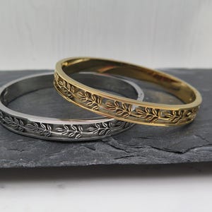 Könnte beinhalten: Zwei dekorative Armreifen, einer silberfarben und einer goldfarben, liegen auf einer dunkelgrauen Oberfläche. Beide Armreifen weisen ein sich wiederholendes Blattmuster um das Band auf. Der goldene Armreif ist leicht erhöht, was seine Details hervorhebt.