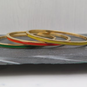 Könnte beinhalten: Drei goldene Armreifen mit einem einzigen farbigen Streifen. Die Armreifen sind übereinander gestapelt. Der obere Armreif hat einen gelben Streifen, der mittlere Armreif einen orangen Streifen und der untere Armreif einen grünen Streifen.