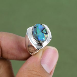 Puede incluir: Un anillo de plata con una incrustación ovalada de concha de abulón. La concha tiene colores iridiscentes azules, verdes y morados.